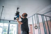 Una persona che indossa un equipaggiamento di sicurezza fora un soffitto utilizzando un martello perforatore a batteria.