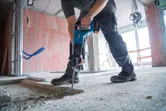 Una persona che indossa un equipaggiamento di sicurezza perfora un pavimento di cemento con un martello perforatore a batteria.