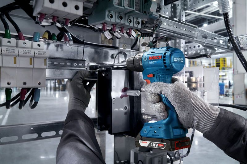 Una persona che indossa un equipaggiamento di sicurezza utilizza un trapano a batteria su un quadro elettrico.