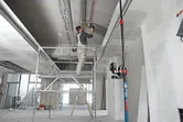 Una persona è in piedi su un'impalcatura e sta installando una conduttura, guidata da uno strumento di livellamento laser su un supporto universale.