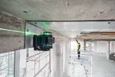 Strumento di livellamento laser che proietta linee verdi sul soffitto, una persona con dispositivi di sicurezza regola l'allineamento.