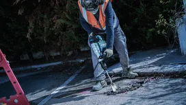 Una persona che indossa l'equipaggiamento di sicurezza rompe l'asfalto con un martello demolitore.