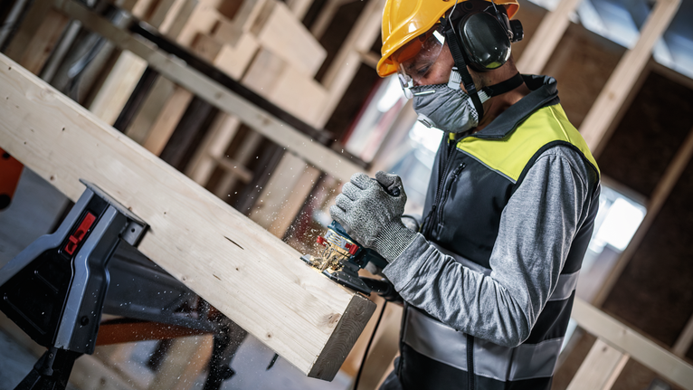 Una persona che indossa un equipaggiamento di sicurezza utilizza un elettroutensile per modellare una trave di legno.