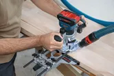 Una persona guida una fresatrice portatile lungo una superficie di legno utilizzando una guida metallica.
