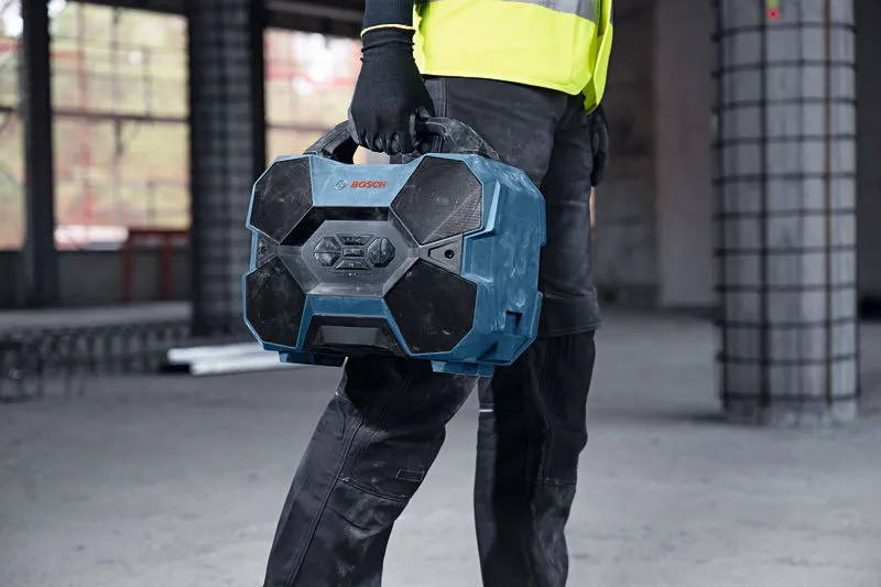 Una persona che indossa un equipaggiamento di sicurezza trasporta una robusta radio blu da cantiere all'interno di un'area di costruzione.