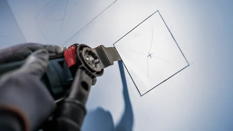 Una persona che indossa un equipaggiamento di sicurezza utilizza un elettroutensile per tagliare un quadrato nel muro a secco.