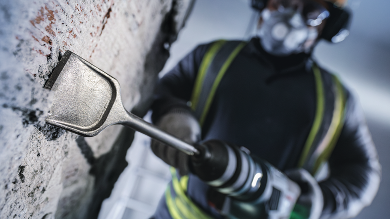 Una persona che indossa un equipaggiamento di sicurezza utilizza un martello perforatore per scalpellare un muro di cemento.