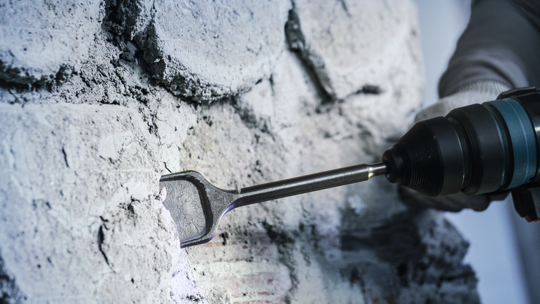 Persona che indossa dispositivi di sicurezza mentre scalpella un muro di cemento con un utensile elettrico.
