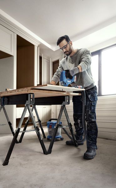 Una persona, munita di dispositivi di protezione individuale, taglia il legno con una sega elettrica su un banco da lavoro.