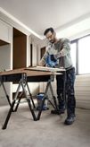 Una persona, munita di dispositivi di protezione individuale, taglia il legno con una sega elettrica su un banco da lavoro.