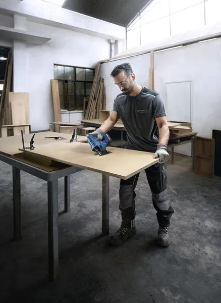 In un'officina, una persona con indosso dispositivi di protezione individuale utilizza una sega elettrica per tagliare una tavola di legno.