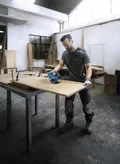In un'officina, una persona con indosso dispositivi di protezione individuale utilizza una sega elettrica per tagliare una tavola di legno.