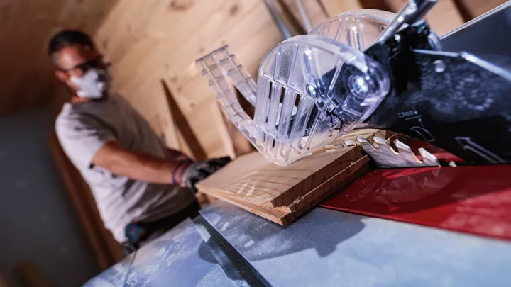 Una persona che indossa dispositivi di sicurezza taglia un'asse di legno con una sega da tavolo.