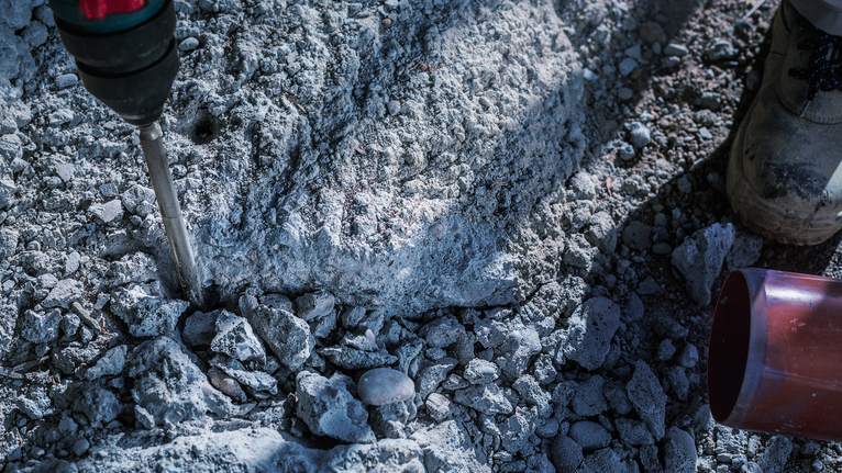 Una persona che indossa un equipaggiamento di sicurezza utilizza un trapano demolitore su calcestruzzo grezzo.