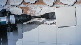Una persona che indossa l'equipaggiamento di sicurezza rimuove le piastrelle del muro con uno scalpello elettrico.