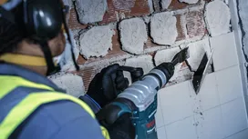Una persona che indossa l'equipaggiamento di sicurezza rimuove le piastrelle del muro con uno scalpello elettrico.