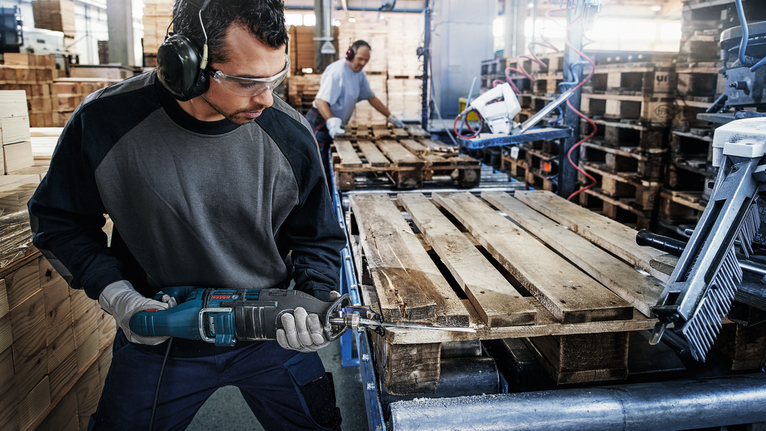Una persona che indossa un equipaggiamento di sicurezza utilizza un elettroutensile per tagliare un pallet di legno.