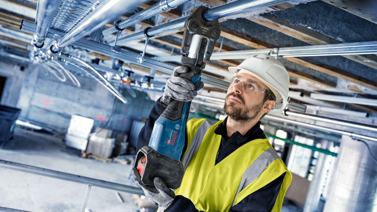 Una persona che indossa un equipaggiamento di sicurezza utilizza un elettroutensile per fissare tubi metallici sopraelevati.