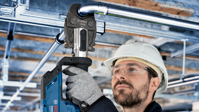 Una persona che indossa un equipaggiamento di sicurezza utilizza un elettroutensile per premere un raccordo per tubi sopra la testa.