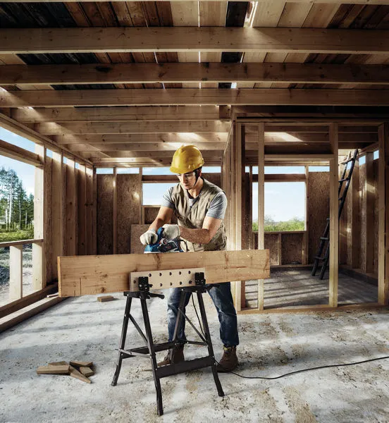 Una persona che indossa un equipaggiamento di sicurezza utilizza una pialla per levigare una trave di legno nella struttura di un edificio.