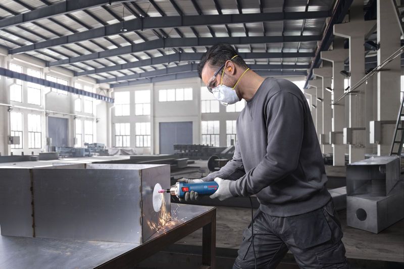 Una persona che indossa dispositivi di sicurezza utilizza una smerigliatrice dritta per levigare una scatola di metallo.