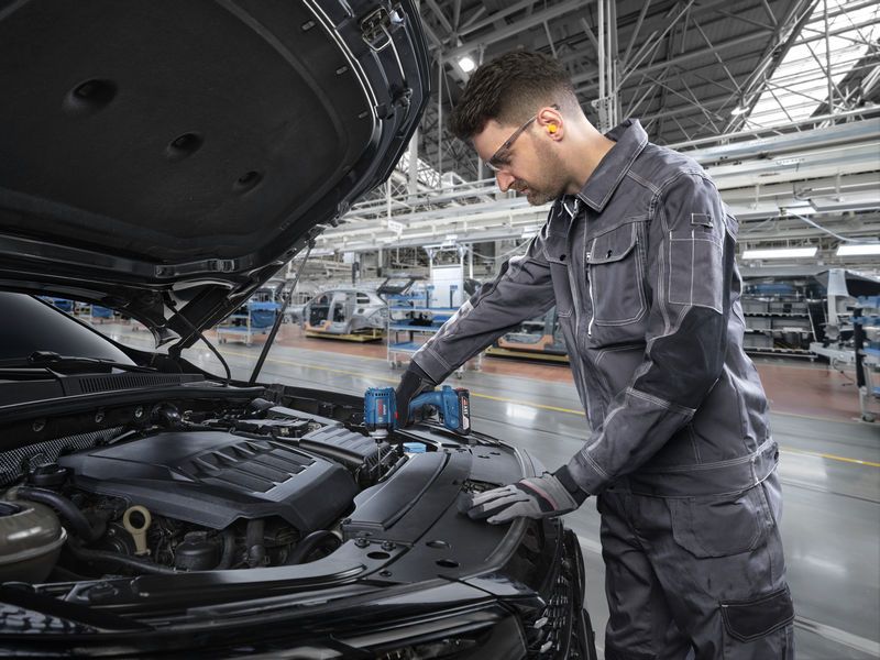 Una persona che indossa un equipaggiamento di sicurezza utilizza un utensile senza fili sul motore di un'auto in una fabbrica.