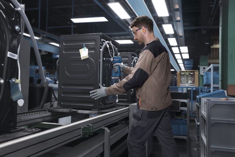 Una persona che indossa un equipaggiamento di sicurezza fissa un elettrodomestico su una catena di montaggio utilizzando un utensile elettrico.