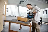 Una persona che indossa dispositivi di sicurezza utilizza una sparachiodi senza fili su una scatola di legno in un'officina.