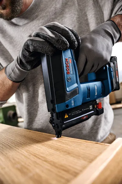 Una persona che indossa un equipaggiamento di sicurezza utilizza una sparachiodi senza fili su una superficie di legno.