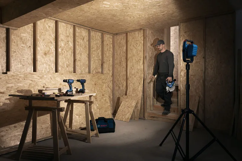 Una persona che indossa dispositivi di sicurezza entra in un laboratorio di falegnameria illuminato da una lampada da cantiere senza fili.