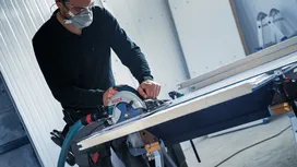 Una persona che indossa dispositivi di sicurezza taglia una tavola con una sega circolare su un tavolo da lavoro.