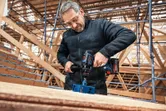 Una persona che indossa un equipaggiamento di sicurezza pratica un grande foro nel legno con un trapano a batteria.