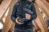 Una persona tiene in mano un trapano a batteria e una batteria all'interno di una struttura in legno.