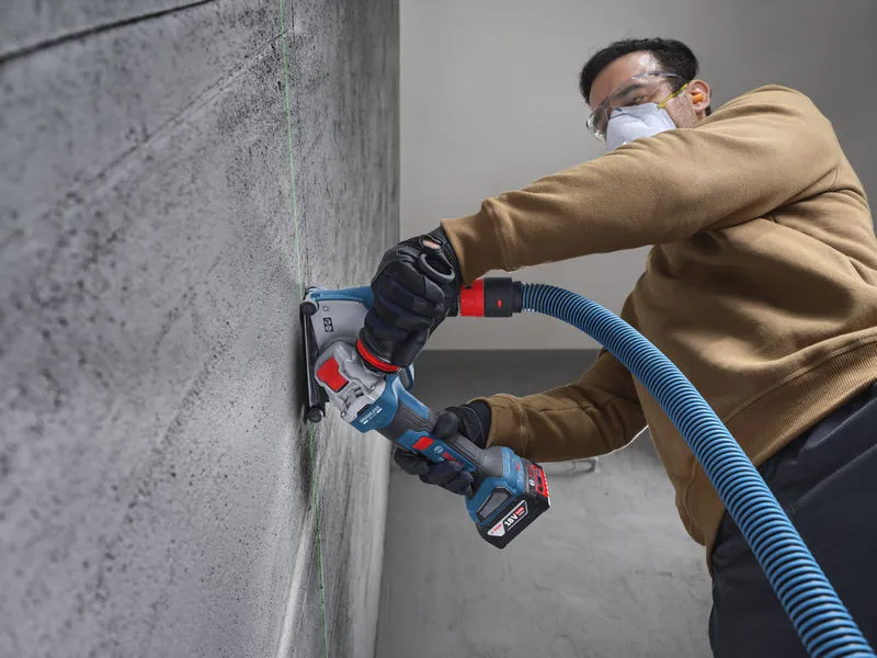 Una persona che indossa un equipaggiamento di sicurezza leviga un muro di cemento con una smerigliatrice angolare a batteria.