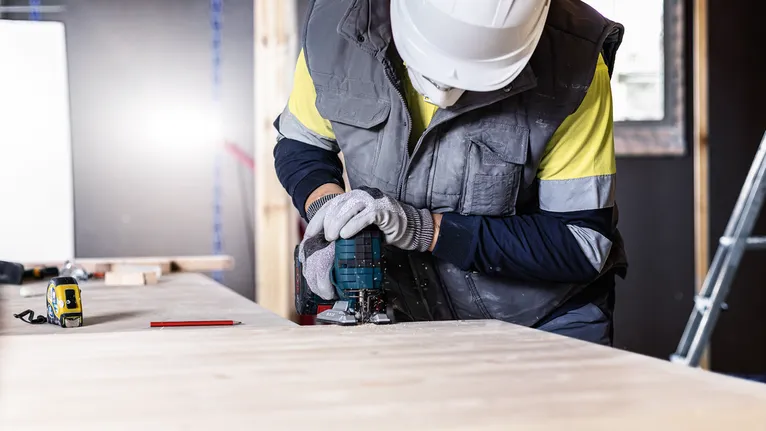 Una persona che indossa dispositivi di sicurezza utilizza un elettroutensile per tagliare la legna su un banco da lavoro.