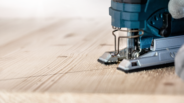 Una persona guida un seghetto alternativo lungo una linea tracciata, tagliando un'asse di legno.