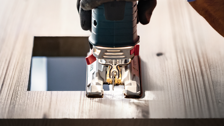 Una persona che indossa dispositivi di sicurezza utilizza un seghetto alternativo per tagliare un'apertura quadrata nel legno.