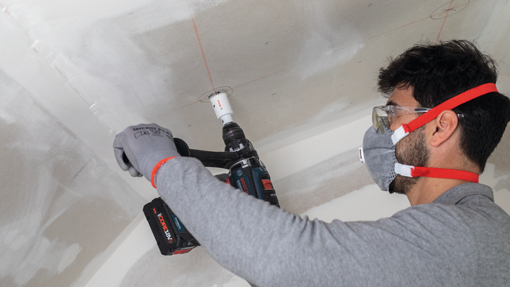Una persona che indossa un equipaggiamento di sicurezza pratica un foro nel soffitto con un utensile elettrico.