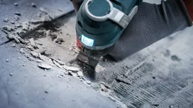 Persona che indossa dispositivi di sicurezza e utilizza un utensile multiuso per rimuovere del materiale dal pavimento.