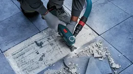 Una persona che indossa dispositivi di sicurezza rimuove l'adesivo per piastrelle con un elettroutensile su un pavimento piastrellato.