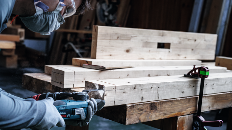 Una persona che indossa un equipaggiamento di sicurezza leviga il bordo di una trave di legno.