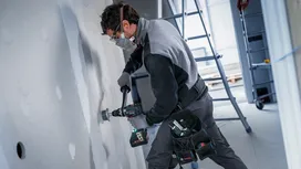 Una persona che indossa un equipaggiamento di sicurezza fora il muro a secco con un utensile elettrico.