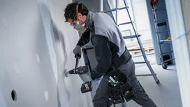 Una persona che indossa dispositivi di sicurezza pratica un foro nel muro a secco in un cantiere edile.