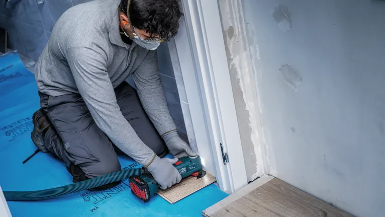 Una persona che indossa dispositivi di sicurezza utilizza un elettroutensile per tagliare il pavimento vicino a una porta.