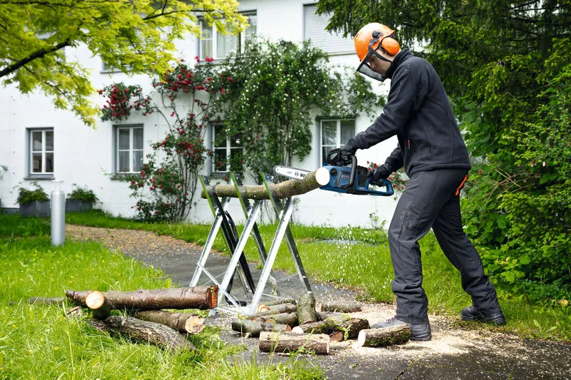 Una persona, munita di dispositivi di protezione individuale, utilizza una motosega a batteria per tagliare i rami di un albero all'aperto.