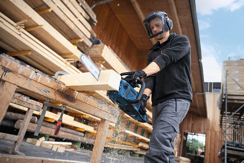 Una persona, munita di dispositivi di protezione individuale, taglia una trave di legno con una motosega a batteria.