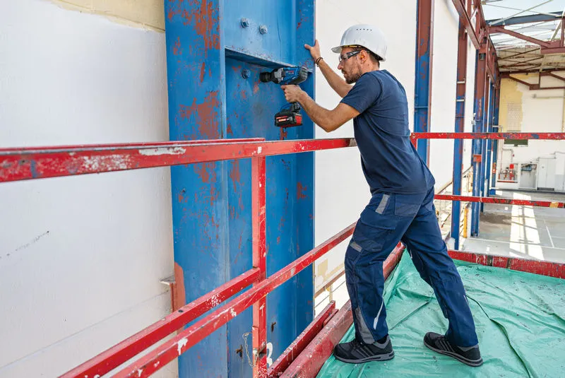 Una persona che indossa un equipaggiamento di sicurezza utilizza una chiave a impulsi senza fili su una trave d'acciaio.