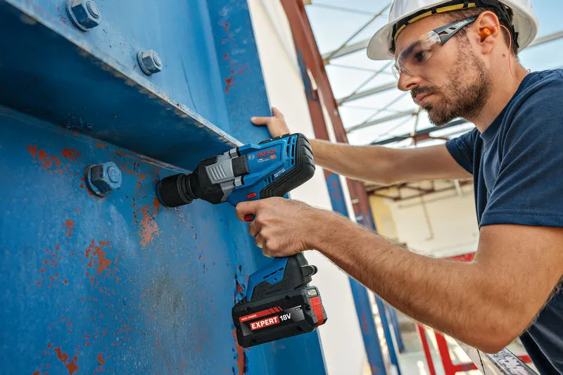 Una persona che indossa un equipaggiamento di sicurezza utilizza una chiave a impulsi senza fili su grandi travi di acciaio blu.