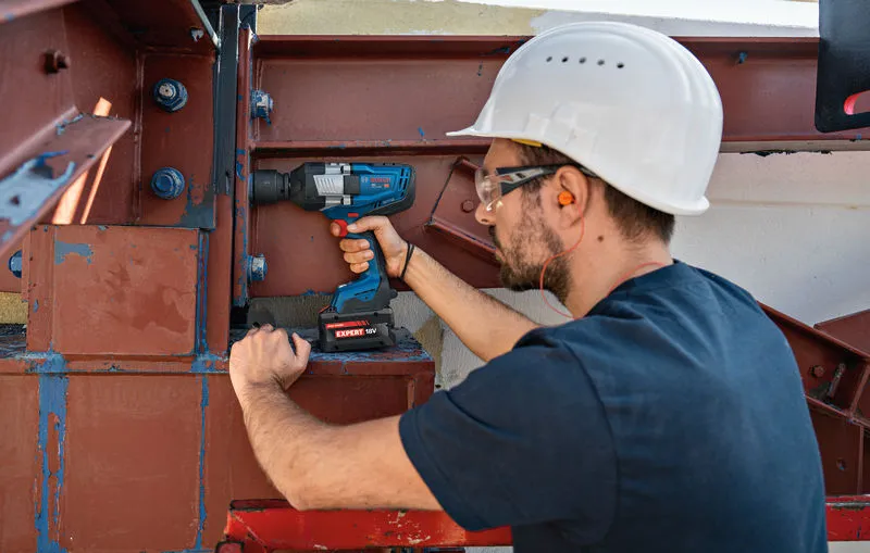 Una persona che indossa un equipaggiamento di sicurezza utilizza una chiave a impulsi senza fili su una trave d'acciaio.