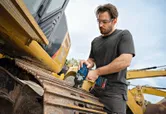 Una persona che indossa un equipaggiamento di sicurezza utilizza una chiave a impulsi a batteria sui cingoli di un escavatore.